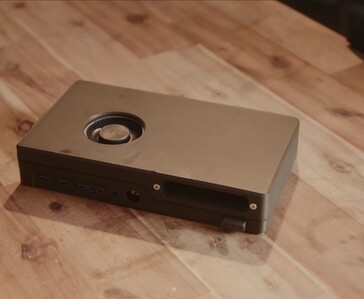 A compact black game console with a built-in disc drive, USB ports and top-mounted cooling fan resting on a wooden table.