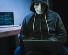 A masked, hooded figure works on a laptop in a darkened room.