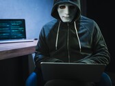 A masked, hooded figure works on a laptop in a darkened room.