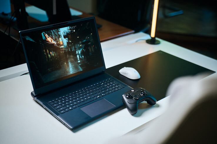 An open Alienware gaming laptop set up on a white studio desk running a dark, reflective street scene in sharp path-traced detail, paired with a black controller and white wireless mouse.