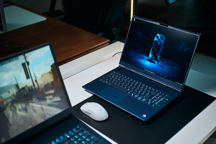 A dark blue mid-range gaming laptop (Alienware Aurora 16X) angled on a desk pad displaying a glowing blue sci-fi wallpaper, positioned next to a white wireless mouse with another laptop blurred in the foreground.