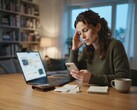 A woman at a desk looks at her smartphone; next to it are a laptop, receipts, and a notepad
