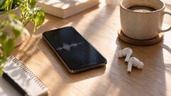 Smartphone displaying an audio waveform beside wireless earbuds and a coffee cup on a wooden desk
