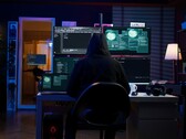 A hooded researcher works across multiple screens in a darkened setup. Generic hacker pictured. 