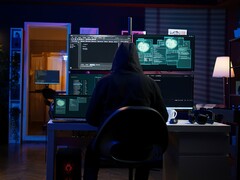A hooded researcher works across multiple screens in a darkened setup. Generic hacker pictured.