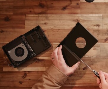 A person disassembling a small black game console on a wooden workbench, with the console casing removed and its internal cooling fan and circuit board visible.