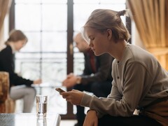 A teenager looks at her smartphone, with two adults sitting out of focus in the background.