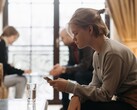 A teenager looks at her smartphone, with two adults sitting out of focus in the background.