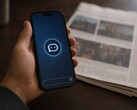 A hand holds a smartphone showing a chatbot icon, a printed newspaper lies next to it.