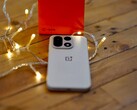 A OnePlus 15 lies on a wooden table surrounded by warm string lights, with a bright red OnePlus retail box positioned in the background.