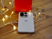 A OnePlus 15 lies on a wooden table surrounded by warm string lights, with a bright red OnePlus retail box positioned in the background.