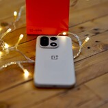 A OnePlus 15 lies on a wooden table surrounded by warm string lights, with a bright red OnePlus retail box positioned in the background.