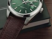 Close-up of the Timex Legacy Day and Date watch, showing its green sunray dial, silver-tone bezel and brushed lugs, ridged crown, and textured brown leather strap against a dark green box and wooden surface.
