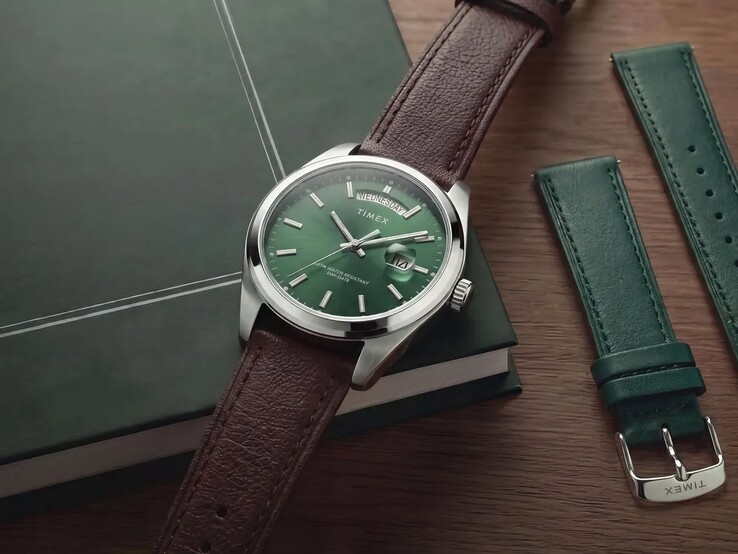 The Timex Legacy Day and Date with its green dial and brown leather strap displayed on a dark green book beside the interchangeable green leather strap.