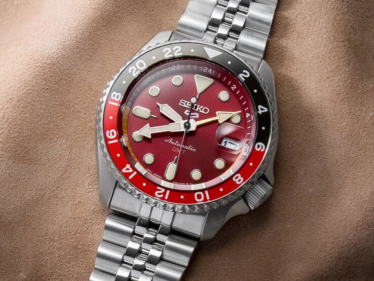 Seiko 5 Sports GMT HDB016 photographed against a brown textured background, showing its burgundy sunray dial, red-and-brown 24-hour bezel, magnified date window, and stainless steel bracelet.