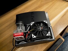 Custom water-cooled PlayStation 3 with a red SysCooling pump and large black fan mounted inside the case, placed on a wooden desk. (Image source: Zac Builds via YouTube)