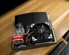 Custom water-cooled PlayStation 3 with a red SysCooling pump and large black fan mounted inside the case, placed on a wooden desk. (Image source: Zac Builds via YouTube)