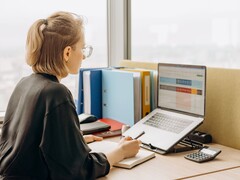 Woman with glasses works on a laptop spreadsheet, with binders and a calculator on the desk