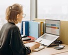 Woman with glasses works on a laptop spreadsheet, with binders and a calculator on the desk