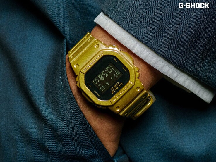 Close-up of a gold Casio G-Shock DW-5600BEP-9 watch worn on a wrist against dark blue fabric. The watch has a black digital display, gold case and band, and BlackEyePatch branding on the bezel and dial.
