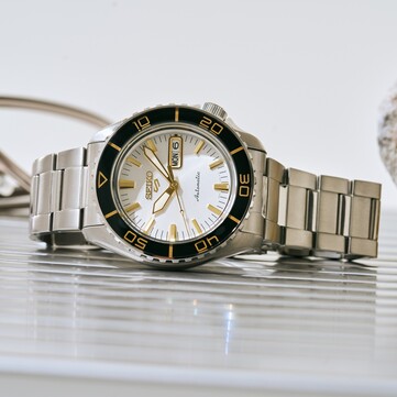 Seiko 5 Sports SKX HDB004 watch with a silver-white dial, black rotating bezel with gold-colored markings, gold-tone hands and indexes, and stainless steel bracelet, photographed at an angle.