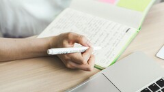 A person is writing notes in a notebook next to an open laptop