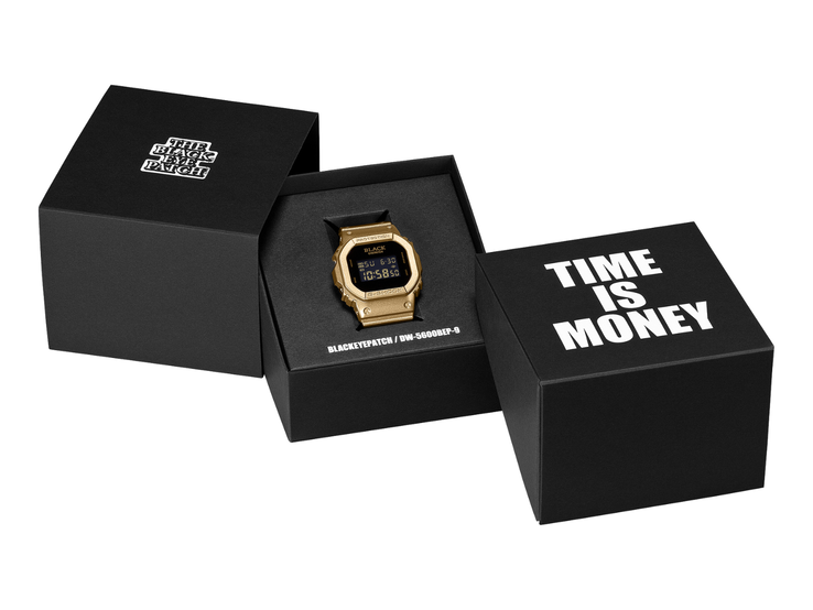 A gold Casio G-Shock DW-5600BEP-9 watch with a black digital display sits between two black presentation boxes. The watch features BlackEyePatch branding on the dial, while the foreground box reads “TIME IS MONEY.”