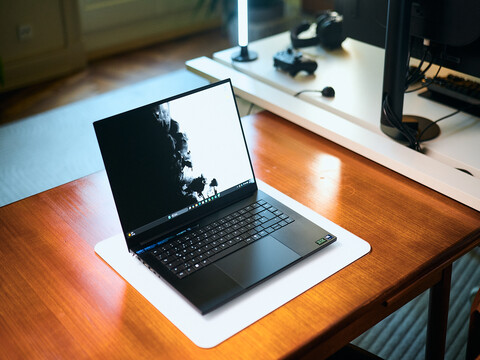 Matte black Razer Blade 16 gaming laptop open on a wooden desk displaying a high-contrast wallpaper, with a desk setup in the background.
