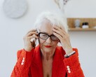 An older woman with white hair and glasses on the phone in a bright living room, holding her hand to her temple.