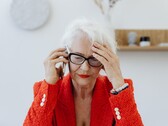 An older woman with white hair and glasses on the phone in a bright living room, holding her hand to her temple.