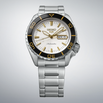 Seiko 5 Sports SKX HDB004 with a silver-white dial, black bezel with gold-colored markings, gold-tone hands and indexes, and stainless steel bracelet shown against a light gray background.